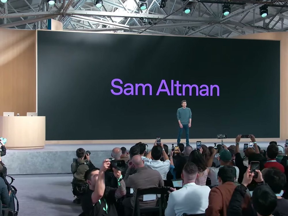 DevDay 2026 舞台：Sam Altman 站在大螢幕前，螢幕上是紫色字的 Sam Altman，台下觀眾舉著手機拍攝