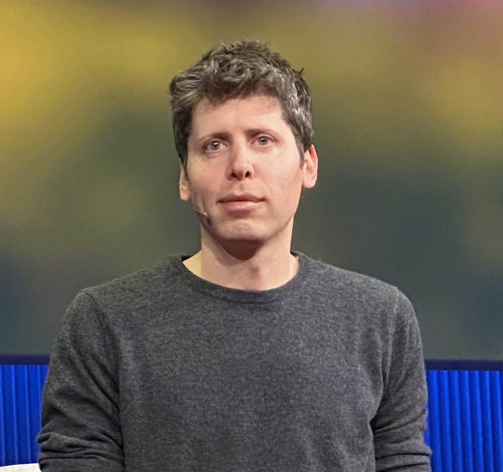 Sam Altman 的半身照，穿深灰色上衣，背景是模糊的黃綠色燈光